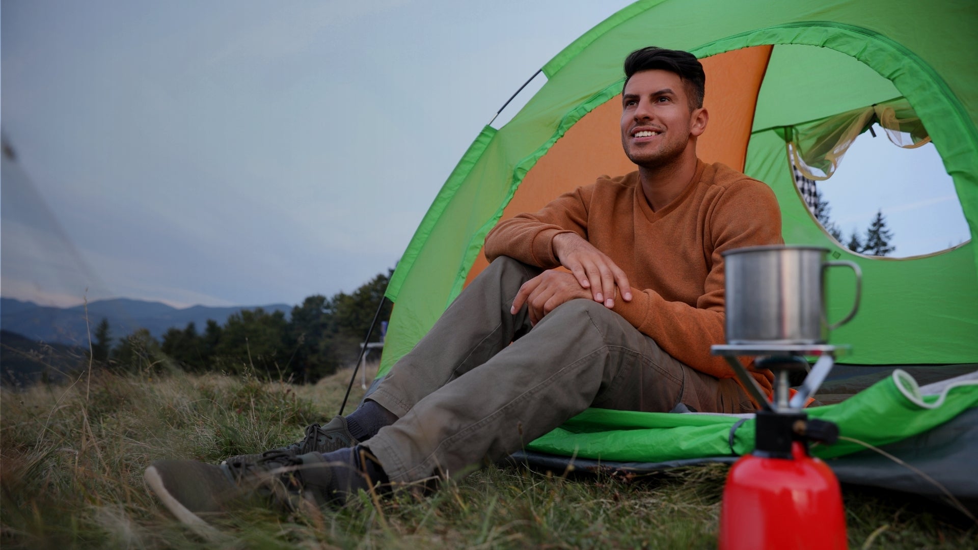 Camper relaxing outside green tent with portable camping stove in mountain meadow